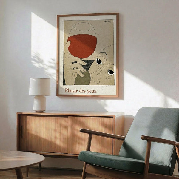 May include: Vintage-style art poster in wooden frame hanging above mid-century modern wood credenza with table lamp. Green upholstered armchair with wooden arms sits nearby. Artwork shows abstract figures with wine glass in muted orange, green and cream tones with French text "Plaisir des yeux" below. Natural sunlight creates geometric shadows on white wall in modern living room setting.