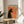 May include: Framed vintage-style poster featuring a black cat silhouette smoking with curled green smoke against orange background. French text reads "Le Chat Nocturne" at top with museum exhibition details below. Poster displayed on round wooden side table next to beige upholstered chair, with plant leaves visible in soft natural lighting.