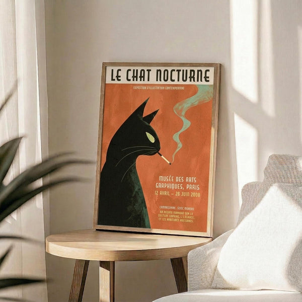 May include: Framed vintage-style poster featuring a black cat silhouette smoking with curled green smoke against orange background. French text reads "Le Chat Nocturne" at top with museum exhibition details below. Poster displayed on round wooden side table next to beige upholstered chair, with plant leaves visible in soft natural lighting.