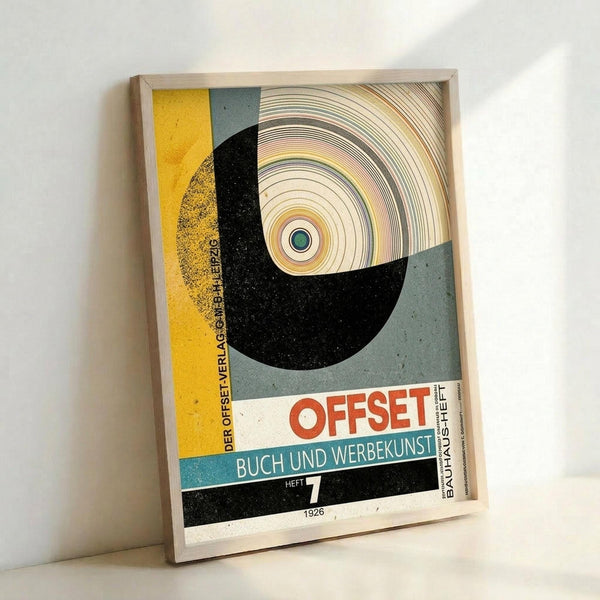 Framed vintage Bauhaus poster featuring bold geometric design with large black semicircle, concentric circles in earth tones, and color blocks in yellow, teal, and gray. German text reads "OFFSET BUCH UND WERBEKUNST HEFT 7 1926" in red and blue lettering. The modernist print has a weathered, aged appearance with speckled texture throughout. Light wooden frame displays the artwork against a white wall corner.