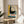 May include: Framed vintage Bauhaus poster with geometric design featuring concentric circles, bold typography reading "OFFSET BUCH UND WERBEKUNST," and color blocks in yellow, green, black and orange. Wood frame sits on round wooden side table next to beige upholstered chair with white throw pillow. Natural sunlight creates shadows on neutral wall behind, with plant leaves visible in corner.