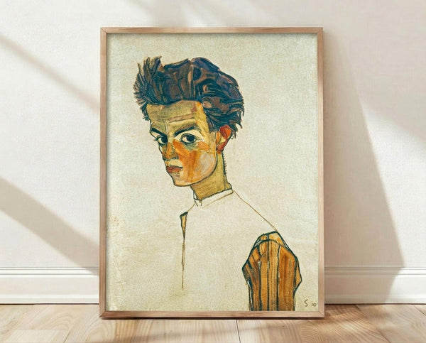 May include: Framed reproduction of an expressionist self-portrait showing a young man with tousled dark hair and angular features, rendered in earthy tones of yellow, orange, and blue against a cream background. The artwork is displayed in a light wood frame, leaning against a white wall on light wooden flooring with soft natural lighting creating subtle shadows.