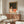 May include: Vintage 1920s poster art by René Vincent displayed in gold ornate frame above wooden sideboard in modern living room. Artwork shows figure in cream robe reading under starry night sky. Room features white curved chair, round glass coffee table, table lamp, and minimalist decor with warm wood tones and neutral palette.