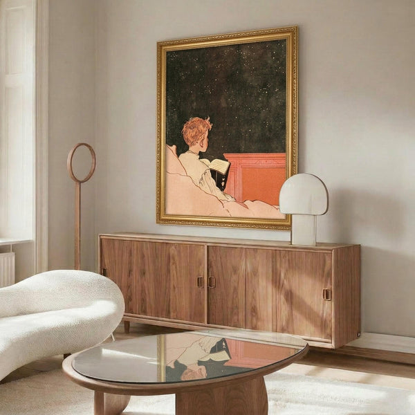 May include: Vintage 1920s poster art by René Vincent displayed in gold ornate frame above wooden sideboard in modern living room. Artwork shows figure in cream robe reading under starry night sky. Room features white curved chair, round glass coffee table, table lamp, and minimalist decor with warm wood tones and neutral palette.