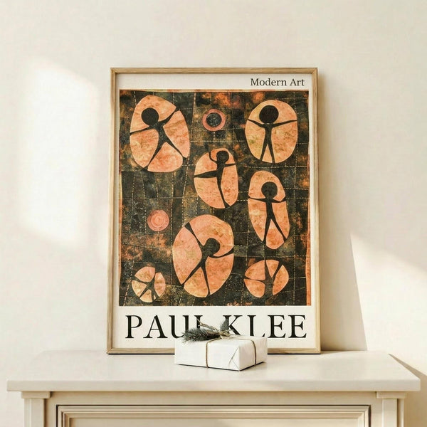 May include: Framed Paul Klee modern art poster displayed on light wood console table against white wall. Abstract artwork features coral pink figures with black stick-like bodies arranged in geometric grid pattern on dark textured background. Small wrapped gift box with greenery sits beside the wooden-framed print. Minimalist home decor styling with natural lighting creating soft shadows.