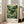 May include: Framed art print displayed in bright living room with beige sofa and large windows. The artwork features rolling green hills and mountains with yellow flowering trees in foreground, decorative sun with radiating lines in sky, and text reading "Yayoi Kusama" and "Voice of the Deep" at bottom. Natural wood frame leans against white wall on hardwood floor.