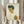 May include: Person in cream dress holding framed reproduction of expressionist self-portrait featuring young man with tousled dark hair, pale complexion, and white collar against beige background. Dried wheat stems visible beside frame in soft natural lighting near window with flowing curtains.