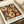 Modern art print featuring abstract human figures in coral and peach tones against a dark textured background with grid pattern. The artwork shows simplified stick-figure forms arranged in circular compositions. Print is matted in cream colored frame, lying flat on white surface next to rolled paper tubes. Artist name "PAUL KLEE" printed at bottom of piece.