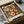 May include: Paul Klee art print in black frame, placed in cardboard shipping box with white tissue paper. The artwork features orange and rust-colored abstract human figures arranged in a grid pattern against a dark weathered background. Wooden dowels and glass bowl visible nearby on neutral surface.