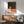 May include: Framed vintage poster showing person with reddish hair in white clothing reading book against starry night sky background in coral and dark green tones. Displayed above wooden sideboard with white ceramic lamp in modern living room with mint green upholstered chair and natural lighting creating geometric shadows on white wall.