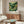 May include: Framed art print featuring rolling green hills, yellow flowering trees, and mountains under a golden sun in Japanese landscape style, displayed above a mid-century walnut credenza with sliding doors. White sculptural chair and oval glass coffee table complete the modern living room setting with cream walls and natural lighting.