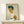 May include: Framed expressionist self-portrait print showing a young man with tousled dark blue hair and angular features in yellow and orange tones, wearing a white collared shirt with orange sleeves. The artwork is displayed in a light wooden frame on a pale wooden shelf against a cream wall, with a small white wrapped gift box placed in front.