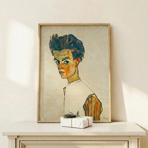 May include: Framed expressionist self-portrait print showing a young man with tousled dark blue hair and angular features in yellow and orange tones, wearing a white collared shirt with orange sleeves. The artwork is displayed in a light wooden frame on a pale wooden shelf against a cream wall, with a small white wrapped gift box placed in front.