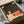 May include: Vintage-style art print in black frame showing person reading under starry night sky, displayed in cardboard shipping box with tissue paper and wooden dowel rods for protection, sitting on white fabric surface next to dark glass object