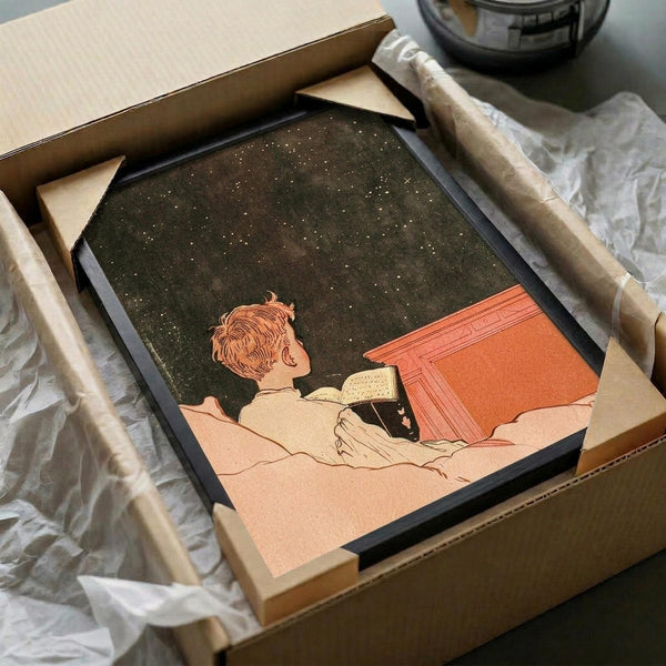 May include: Vintage-style art print in black frame showing person reading under starry night sky, displayed in cardboard shipping box with tissue paper and wooden dowel rods for protection, sitting on white fabric surface next to dark glass object