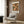 May include: A framed Paul Klee poster featuring abstract orange and black figures on a dark textured background, displayed in a modern living room. The cream-colored armchair has a geometric patterned pillow, positioned next to a brass floor lamp and dark round side table with an open book. Warm wood paneling creates a sophisticated backdrop.