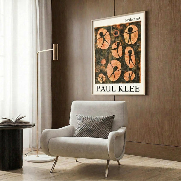 May include: A framed Paul Klee poster featuring abstract orange and black figures on a dark textured background, displayed in a modern living room. The cream-colored armchair has a geometric patterned pillow, positioned next to a brass floor lamp and dark round side table with an open book. Warm wood paneling creates a sophisticated backdrop.