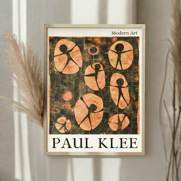 May include: Framed art print featuring abstract stick figures in peachy-orange tones against a dark gridded background, displayed on a light wall next to dried pampas grass stems. The modern artwork shows simplified human forms arranged within circular and oval shapes, with "Modern Art" and "PAUL KLEE" text visible on the print. Light wood frame complements the neutral home decor setting.
