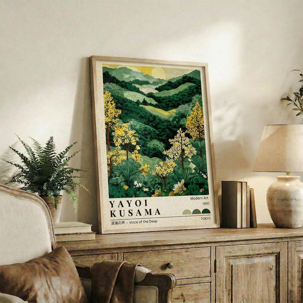 May include: Framed landscape artwork displaying rolling green hills, yellow flowering trees, and mountains under a golden sun, styled as a vintage exhibition poster with "YAYOI KUSAMA" text at bottom. Light wood frame sits on rustic wooden sideboard alongside fern plant, ceramic vases, and books in neutral-toned living room setting.