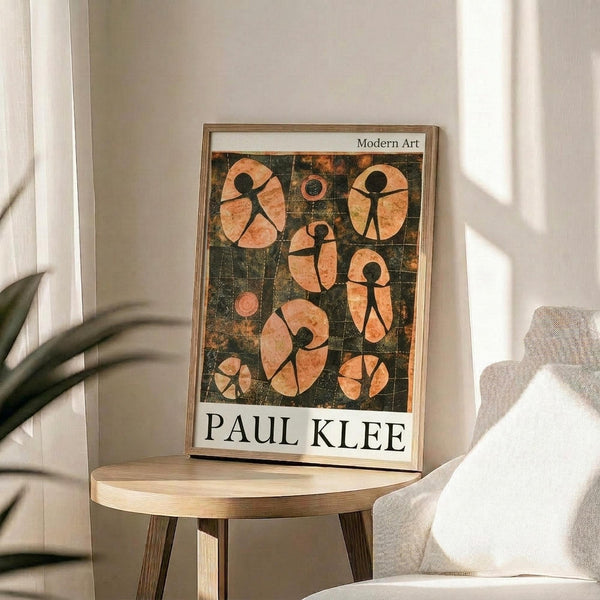 May include: Framed art print featuring abstract figures in orange and peach tones against dark green background, displayed on round wooden side table next to beige upholstered chair. Natural light streams across white walls creating soft shadows. Green plant leaves visible in corner of minimalist interior setting.