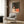 May include: Vintage 1920s art poster showing a person with reddish hair reading in bed while looking out at a starry night sky through a window. The artwork features warm coral and green tones in an Art Deco style. Displayed in a light wood frame on dark wood paneled wall above a beige upholstered armchair with geometric patterned pillow. Modern reading lamp and side table with open book complete the cozy interior setting.