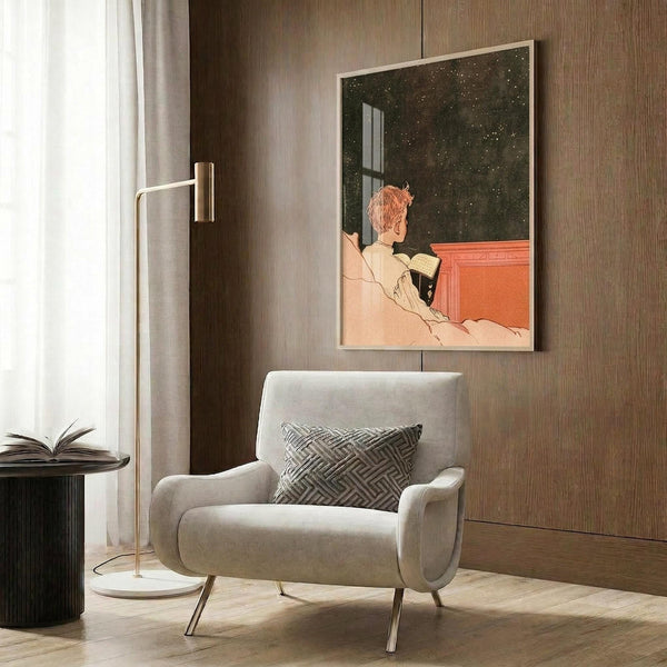 May include: Vintage 1920s art poster showing a person with reddish hair reading in bed while looking out at a starry night sky through a window. The artwork features warm coral and green tones in an Art Deco style. Displayed in a light wood frame on dark wood paneled wall above a beige upholstered armchair with geometric patterned pillow. Modern reading lamp and side table with open book complete the cozy interior setting.