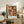 May include: Framed modern art poster featuring abstract figures in coral and peach tones against dark green grid background, displayed on rustic wooden sideboard next to fern plant, ceramic vase, and books in neutral-toned living room setting with beige armchair visible.