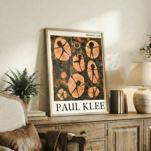 May include: Framed modern art poster featuring abstract figures in coral and peach tones against dark green grid background, displayed on rustic wooden sideboard next to fern plant, ceramic vase, and books in neutral-toned living room setting with beige armchair visible.