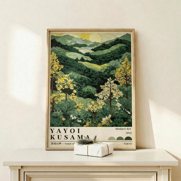 May include: Framed art print displaying rolling green hills and mountains with yellow flowering trees in foreground, pale sun in sky, and text reading "Yayoi Kusama Modern Art 1950 Tokyo" at bottom. Natural wood frame sits on light wooden surface against cream wall, with small white wrapped gift box placed in front.