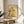 May include: Framed art print featuring botanical illustration with yellow and orange flowers, green foliage, and dotted patterns in Kusama's signature style, displayed on round wooden side table next to beige upholstered chair with white throw pillow, plant visible in corner, natural sunlight streaming across white walls