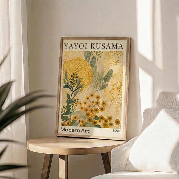 May include: Framed art print featuring botanical illustration with yellow and orange flowers, green foliage, and dotted patterns in Kusama's signature style, displayed on round wooden side table next to beige upholstered chair with white throw pillow, plant visible in corner, natural sunlight streaming across white walls