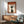 May include: Mid-century modern living room with sage green upholstered armchair and wooden sideboard. Framed poster on white wall shows silhouette of person in profile against cream background with orange geometric squares and Japanese text reading "ZEN" at top. Warm sunlight creates shadows across the space. White ceramic table lamp sits on sideboard next to partial view of round wooden coffee table.