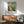 May include: Vintage-style travel poster featuring Navy Pier with Ferris wheel, sailboats on blue water, and classic architecture in warm teal and cream tones. Displayed in thin wood frame above mid-century modern credenza with table lamp, next to sage green upholstered armchair in sunlit room with white walls.