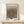 May include: Framed art print with black polka dot pattern radiating from central orange oval, text reading "YAYOI KUSAMA" at top and "Modern A" and "1975" at bottom, displayed on light wood console table with small wrapped gift box beside it against white wall