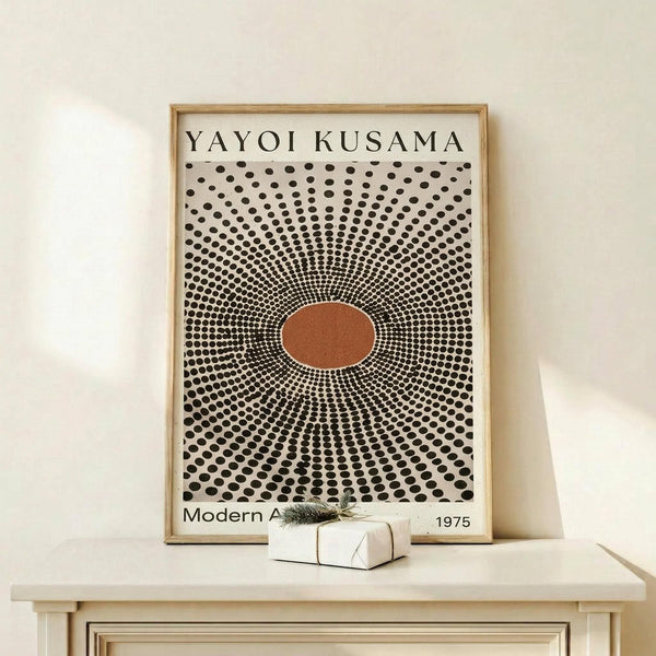 May include: Framed art print with black polka dot pattern radiating from central orange oval, text reading "YAYOI KUSAMA" at top and "Modern A" and "1975" at bottom, displayed on light wood console table with small wrapped gift box beside it against white wall