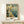 May include: Vintage-style travel poster in wooden frame showing Navy Pier with Ferris wheel, ornate buildings, and sailboats on blue water. Text reads "Welcome to Illinois" and "The Navy Pier" in retro typography. Poster sits on light wood shelf next to small white gift box with evergreen sprig against cream wall.