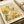 May include: Botanical art print displayed in an open book or portfolio, featuring stylized flowers and foliage in warm yellows, oranges and sage greens with dotted pattern backgrounds. The artwork shows delicate stems, leaves and blooms in a modern illustrative style against cream paper. Text reads "YAYOI KUSAMA" at top, "Modern Art" at bottom, and "1996" in corner.