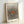May include: Framed art print with Yayoi Kusama's name at top, featuring radiating black polka dots emanating from central orange oval on cream background, with "Modern Art 1975" text below. Light wood frame leaning against white wall, creating subtle shadow. The dots create an optical illusion of movement and depth, transitioning from dense at center to sparse at edges.