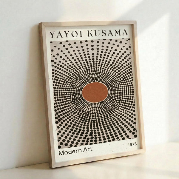 May include: Framed art print with Yayoi Kusama's name at top, featuring radiating black polka dots emanating from central orange oval on cream background, with "Modern Art 1975" text below. Light wood frame leaning against white wall, creating subtle shadow. The dots create an optical illusion of movement and depth, transitioning from dense at center to sparse at edges.