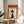 May include: Framed poster with orange and cream design featuring silhouette of person in profile, Japanese text including "ZEN" at top, displayed on round wooden side table next to beige upholstered chair with white throw pillow, plant leaves visible in corner, natural lighting creating soft shadows on white wall