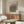 May include: Modern living room with cream walls featuring framed poster with radiating black polka dot pattern on white background with orange center circle. Poster displays "YAYOI KUSAMA" and "Modern Art 1975" text. Room shows walnut wood sideboard, white curved chair, glass coffee table, brass floor lamp, and natural lighting creating warm shadows across wooden furniture.