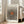 May include: Framed art print with cream background featuring radiating black polka dots in circular pattern around orange-brown center oval, text reading "YAYOI KUSAMA" at top and "Modern Art 1975" at bottom, displayed in light wood frame leaning against white wall next to beige upholstered chair in bright, minimalist living room with large windows and natural lighting.