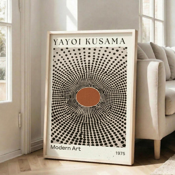 May include: Framed art print with cream background featuring radiating black polka dots in circular pattern around orange-brown center oval, text reading "YAYOI KUSAMA" at top and "Modern Art 1975" at bottom, displayed in light wood frame leaning against white wall next to beige upholstered chair in bright, minimalist living room with large windows and natural lighting.