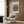 May include: Modern reading corner with beige armchair, geometric throw pillow, and brass floor lamp. Framed poster on wood-paneled wall features radiating black dots forming sunburst pattern around orange center circle. Contemporary interior styling with neutral tones and mid-century furniture elements.