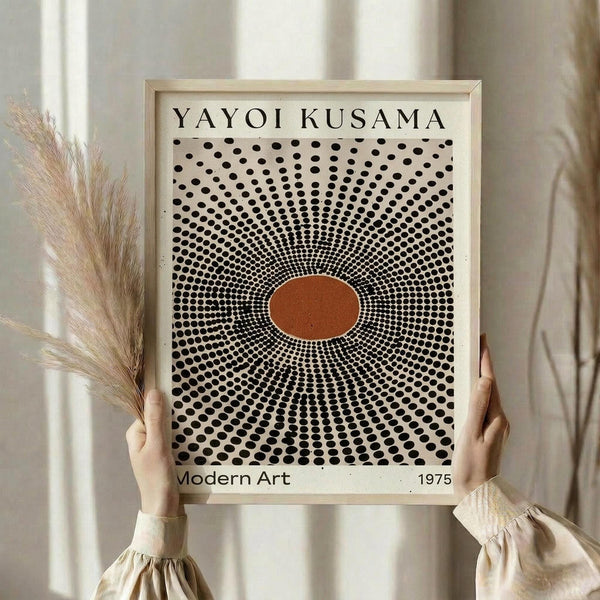 May include: Hands holding a framed art print featuring concentric black polka dots radiating outward from an orange center circle, with "YAYOI KUSAMA" and "Modern Art 1975" text. Light wood frame against cream curtains with dried pampas grass visible on left side. Person wearing beige ruffle-sleeved top.