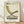 May include: A framed vintage-style poster featuring an olive green mid-century modern lounge chair with tufted upholstery and curved X-base legs. The beige textured background displays "1983" and "Timeless Design" at the top, "VISIONS" in large brown letters below the chair, with smaller descriptive text about form and function. The wooden frame sits on light wood flooring against a white textured wall with soft natural shadows.