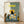 May include: Framed poster print leaning against white wall on light wood floor. Artwork shows black cat sitting at breakfast table with checkered blue tablecloth, yellow chair, plates of eggs and cookies, orange juice bottle, coffee cup. Yellow "CHOW!" text at top, painted in folk art style with green and cream background.