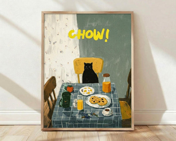 May include: Framed poster print leaning against white wall on light wood floor. Artwork shows black cat sitting at breakfast table with checkered blue tablecloth, yellow chair, plates of eggs and cookies, orange juice bottle, coffee cup. Yellow "CHOW!" text at top, painted in folk art style with green and cream background.