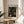 May include: Framed art print featuring black background with golden yellow dotted circular patterns in concentric rings and spirals, displayed on light wood round side table next to beige upholstered chair. Natural lighting creates soft shadows on cream colored wall. Green plant leaves visible in corner. Modern minimalist home decor setting with neutral tones.