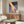 May include: Modern living room with walnut credenza displaying framed poster showing golden hand touching blue water with ripples, creating concentric circles. White sculptural chair and oval glass coffee table in foreground. Warm wood tones complement blue and orange artwork colors. Contemporary minimalist interior styling with natural lighting.